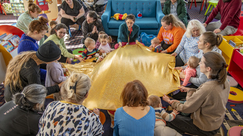 Large group of people holding a gold parachute sitting on a carpeted floor within a library playing a game. Sofas and bookshelves can be seen in the background.