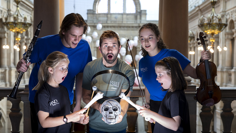 Group of four young people and one adult with surprised expressions, holding a skull, bones, and musical instruments.