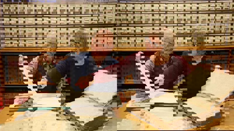 Two people sitting at a desk with two large old books in front of them with filing cabinets behind them in the background