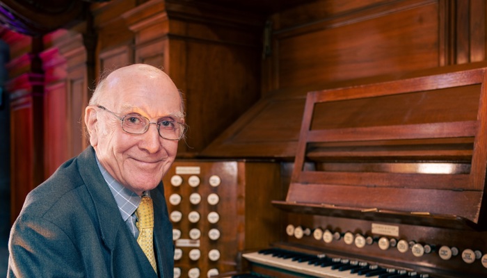 Bill sits at the organ in Kelvingrove Museum smiling at the camera, he is wearing glasses and a grey suit