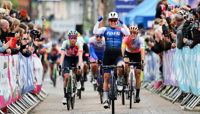 Female cyclists are cheered on by spectators as the lead cyclist celebrates by raising both of her hands in the air