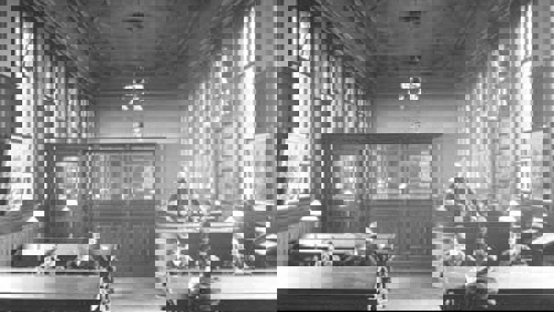 Black and white image showing an old fashioned library, there are large wooden desks and people sitted at them reading books and newspapers