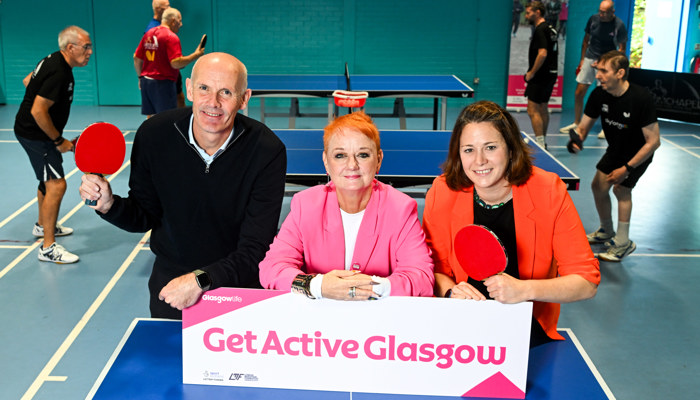 A man and two women hold a sign on a table tennis table which reads 'Get Active Glasgow' while a table tennis match takes place in the background
