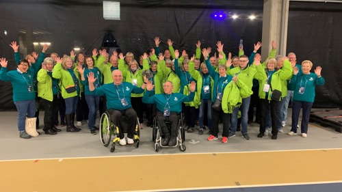 A group of people standing dressed in green smiling with hands in the air, two wheelchair uses are at the front of the group