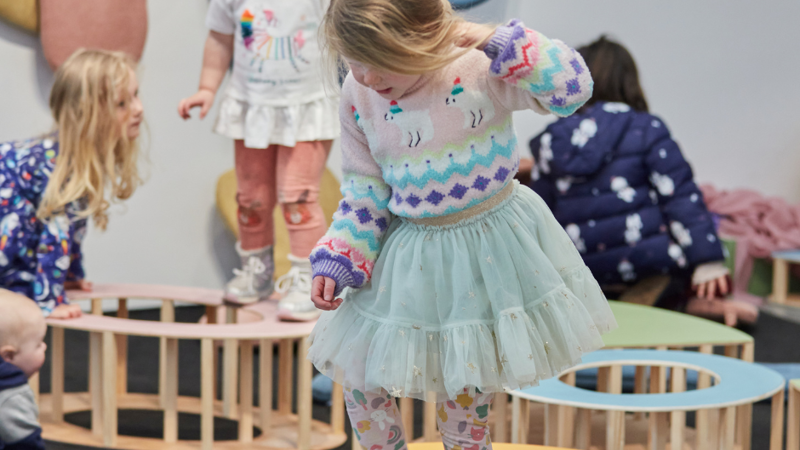  A child in a sweater and skirt steps on colorful, low wooden platforms during interactive exhibition.