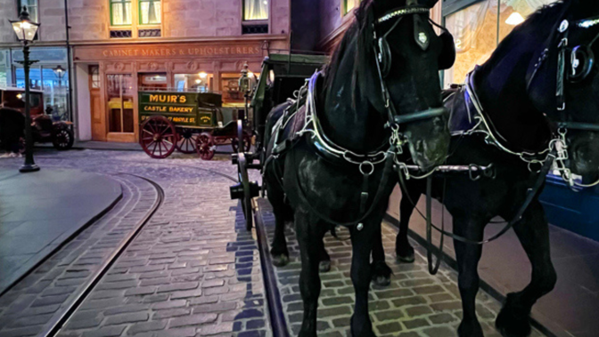 Two dark horses pulling a historic carriage on a cobblestone street in Riverside Museum's indoor period street scene.