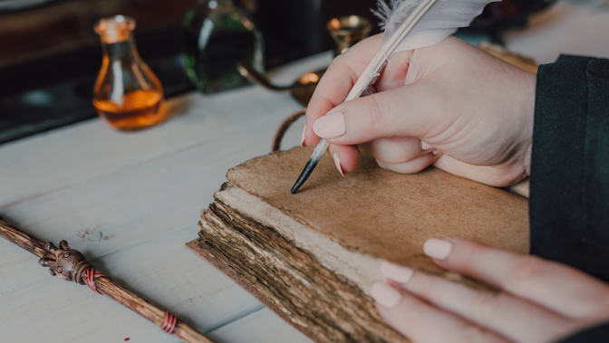  Close-up of a hand with light pink nail polish writing on a worn, leather-bound book with a white feather quill.